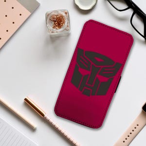 Może przedstawiać: Czerwone etui na telefon z czarnym logo Transformers. Logo przedstawia stylizowaną twarz robota na czarnym tle.