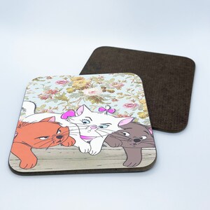 May include: Three cartoon cats on a coaster. The cats are a white cat with a pink bow, an orange cat, and a gray cat. The coaster has a floral pattern and a wood-like texture.