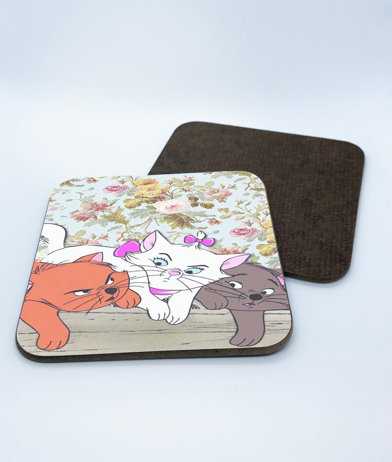 May include: Three cartoon cats on a coaster. The cats are a white cat with a pink bow, an orange cat, and a gray cat. The coaster has a floral pattern and a wood-like texture.