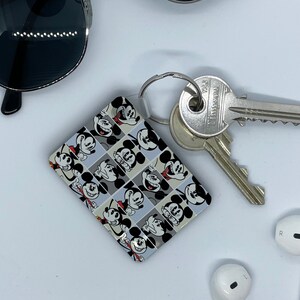 May include: A Mickey Mouse-themed keychain featuring a repeating pattern of Mickey's face in black, white, and red. The keychain is attached to a silver key ring, with two keys and a pair of sunglasses nearby. White earbuds are also visible.