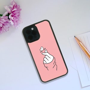 May include: A pink phone case with a black border and a white outline of a hand making a heart sign. The case has a camera cutout and a white heart above the hand.