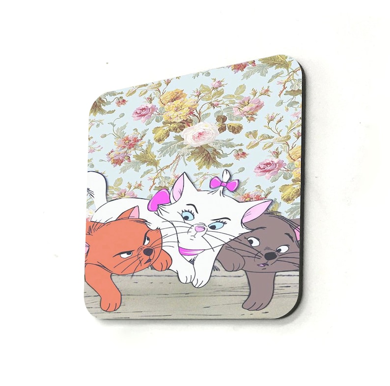 May include: Three cartoon cats, a white cat with a pink bow, a brown cat, and a gray cat, are sitting on a wooden surface with a floral wallpaper background.
