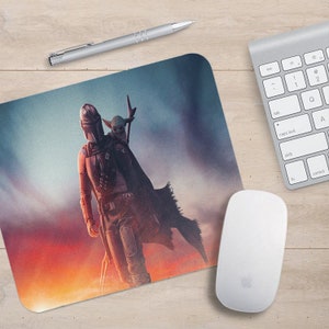 May include: A mouse mat featuring a digital illustration of a Mandalorian character from the Star Wars franchise. The character is wearing a helmet and armour, and is holding a small, green creature. The background is a colourful sunset.