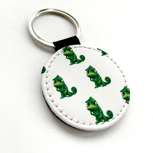 May include: A white round leather keychain with a black leather strap and a silver metal ring. The keychain features a repeating pattern of a green cartoon chameleon with a grumpy expression.