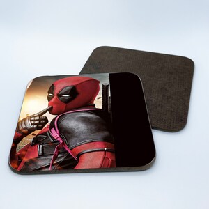 May include: A square coaster featuring a red and black superhero character with a finger over his mouth. The background is a blurry cityscape with a sunset.