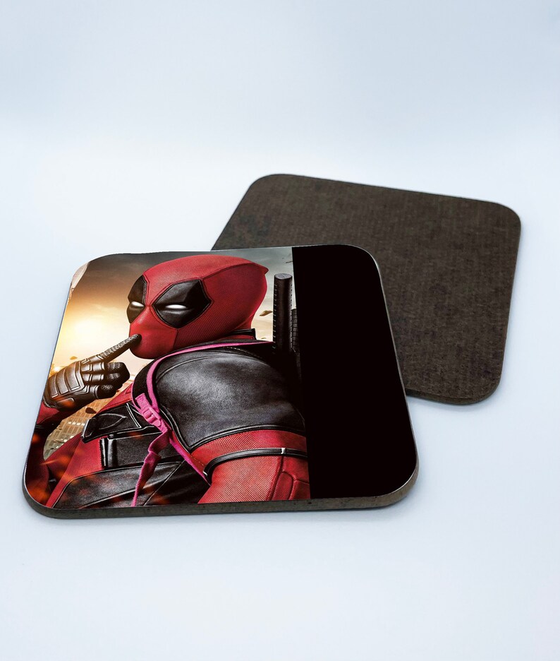 May include: A square coaster featuring a red and black superhero character with a finger over his mouth. The background is a blurry cityscape with a sunset.