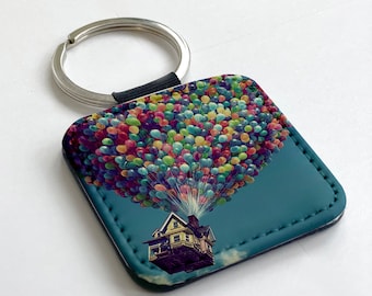 May include: Square keychain featuring a house lifted by colorful balloons against a blue sky. The keychain has a silver ring and a black strap. The image is a whimsical illustration.