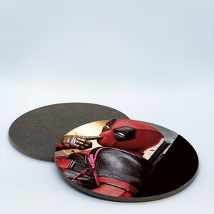 May include: A round coaster with a black background and a printed image of Deadpool in a red and black suit. Deadpool is looking to the right and has his finger to his lips.