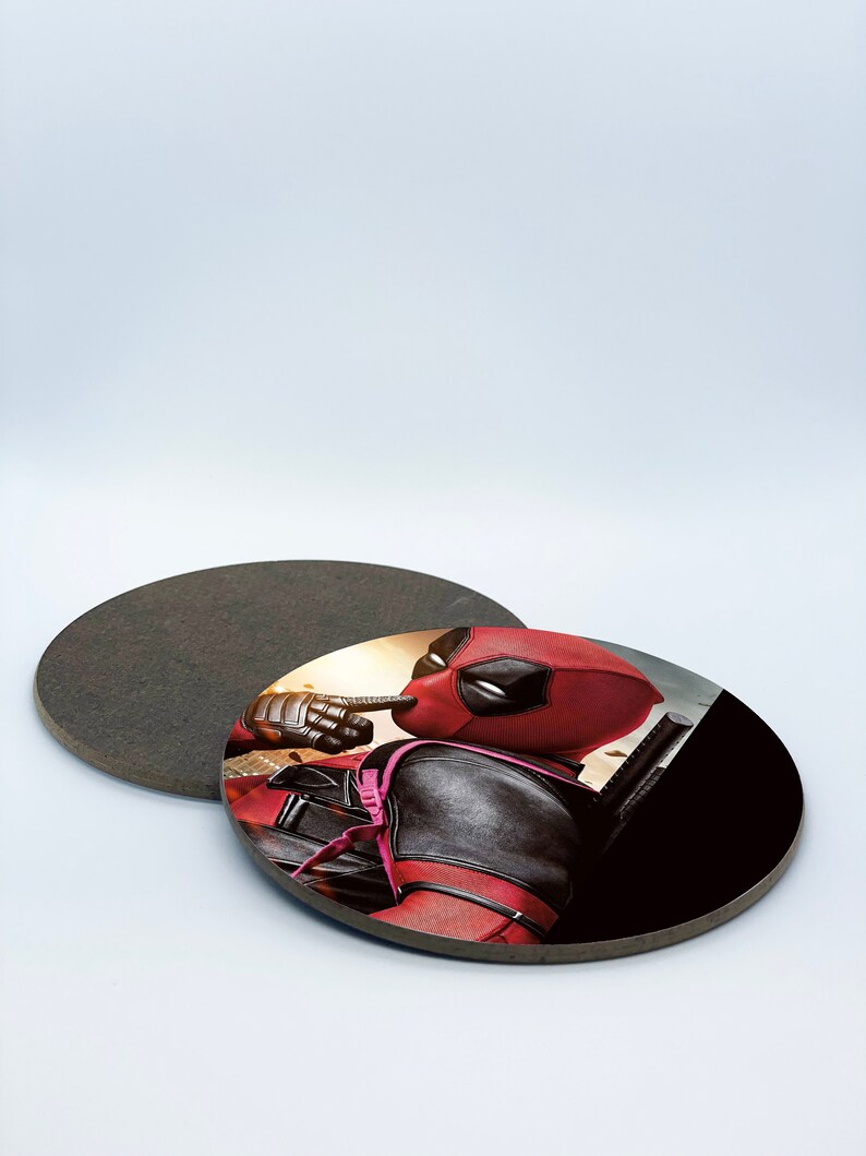 May include: A round coaster with a black background and a printed image of Deadpool in a red and black suit. Deadpool is looking to the right and has his finger to his lips.