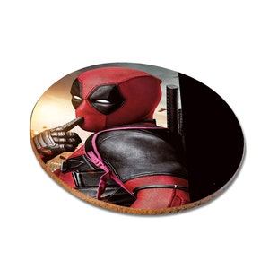 May include: A round coaster featuring a close-up image of Deadpool, a superhero character, wearing his signature red and black suit. He is holding his finger to his lips, as if to tell someone to be quiet. The background is a blurred image of a city skyline.