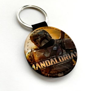 May include: A round leather keychain featuring a character from the Star Wars series, The Mandalorian. The character is wearing a helmet and armor, and the text "The Mandalorian" is visible in the background.