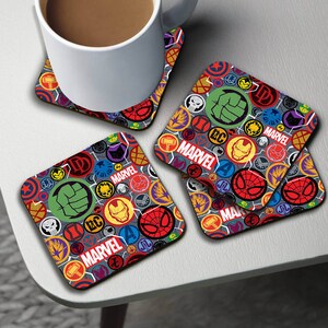 May include: Set of four square coasters featuring a colorful pattern of Marvel superhero logos and symbols on a gray background. The coasters have a wood-like finish and the word "MARVEL" is prominently displayed on each coaster.