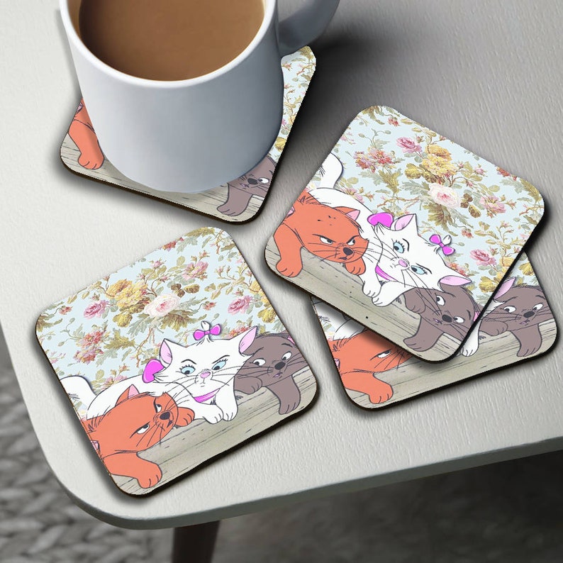 May include: Set of four square coasters featuring a floral pattern with three cartoon cats. The cats are orange, white, and gray and are lying on a wooden surface.
