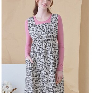May include: A white apron with a black floral pattern. The apron has a square neckline and two pockets. The model is wearing a pink long-sleeved shirt under the apron.