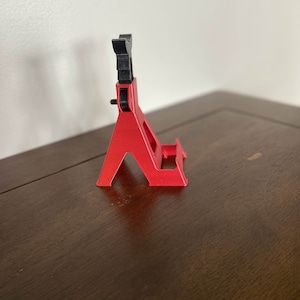 May include: A red plastic phone stand with a black adjustable top. The stand is designed to hold a phone upright.