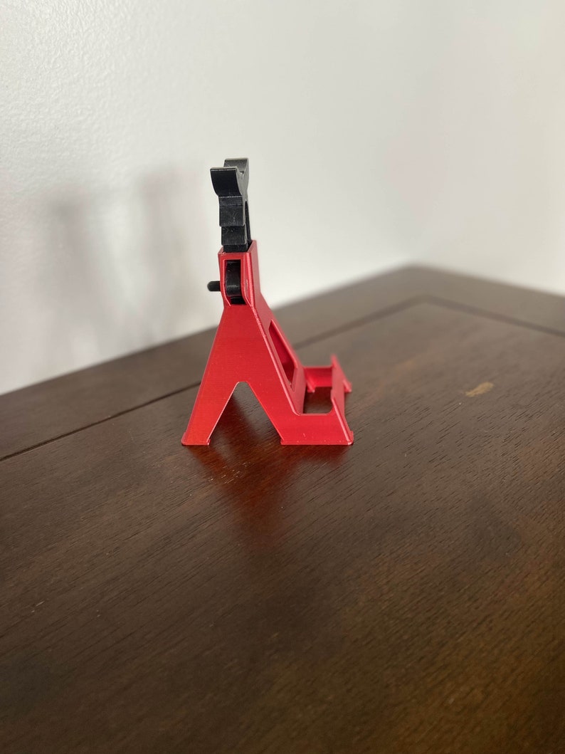 May include: A red plastic phone stand with a black adjustable top. The stand is designed to hold a phone upright.