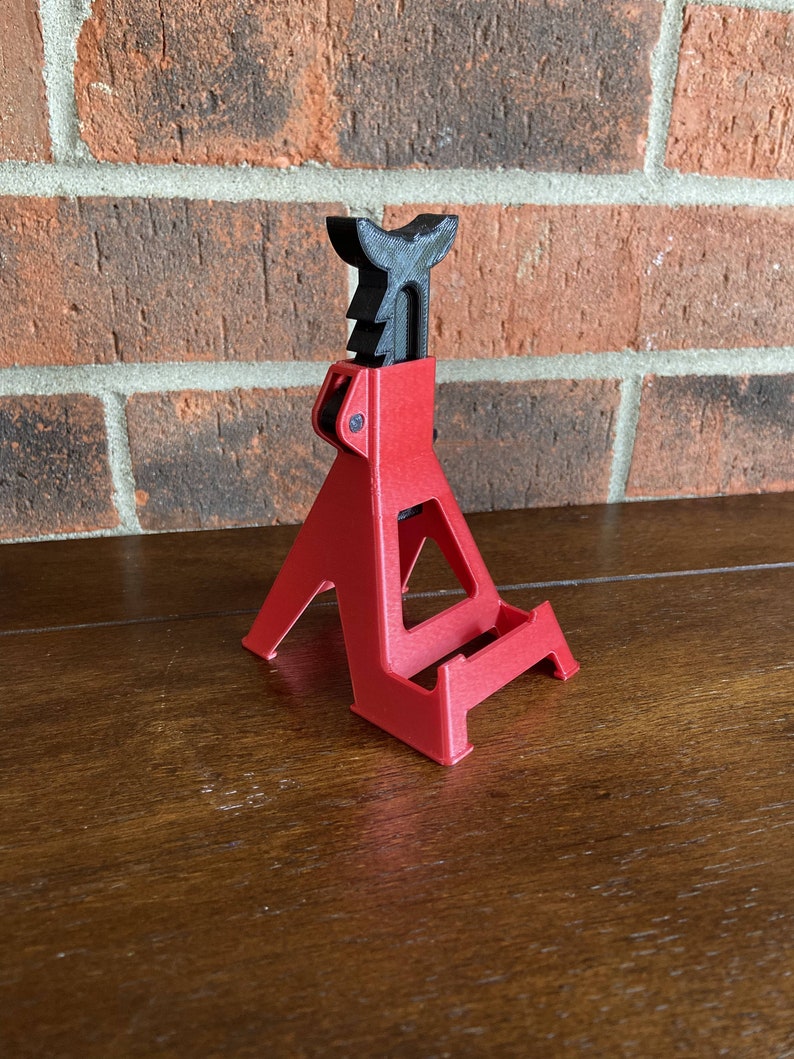 May include: A red plastic phone stand shaped like a car jack. The stand has a black plastic handle and a textured surface.