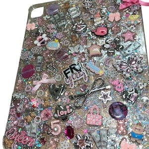 May include: A clear tablet case decorated with a dense arrangement of pink and silver charms, including miniature bows, hearts, flowers, butterflies, teddy bears, dollar bills, and the text "FRI yay" and "LOVE" in a glittery resin finish.