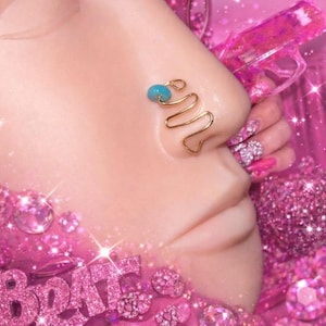 May include: A gold-colored nose cuff with a turquoise bead accent. The cuff has a wavy design and is worn on the nose. The image has a pink background with glitter and other pink accessories.