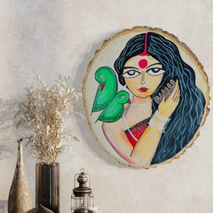 May include: A round wooden wall art piece with a painting of a woman with long black hair, a red bindi, and green birds. The woman is holding a comb in her hand and is looking to the side. The background is a light brown color.