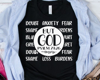 Puede incluir: Camiseta negra con texto blanco que dice "Duda Ansiedad Miedo Vergüenza Pero Dios Salmo 73:26 Duda Vergüenza Pérdida Cargas". La camiseta está doblada y colocada sobre una superficie de madera blanca con una camisa a rayas azul y blanca, una cartera rosa y zapatos de estampado de leopardo.