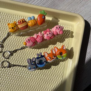 May include: A collection of colorful keychains featuring cartoon characters. The keychains include a yellow bear, a honey pot, a bee, a carrot, pink characters, a bunny, donuts, and foxes. Each keychain has a silver chain and clasp.