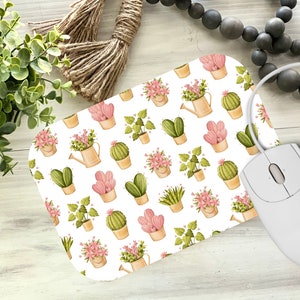 May include: A rectangular mousepad with a white background featuring a repeating pattern of potted plants, cacti, and watering cans in shades of pink, green, and tan. A white computer mouse is connected to the mousepad.
