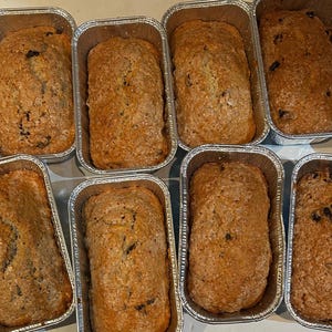 May include: A close-up of nine loaves of banana bread in individual aluminum baking pans. The loaves are all a light brown color and have a slightly crispy crust.