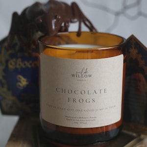 May include: A brown glass candle jar with a white label that reads "wilde WILLOW CANDLE CO. CHOCOLATE FROGS THEY'VE ONLY GOT ONE GOOD JUMP IN THEM Hand poured in Melbourne, Australia Natural soy wax & eco wood wick 300g / 50 hours"