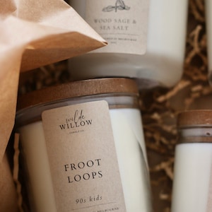 Op de afbeelding: Drie glazen kaarsenpotten met houten deksels en kraftpapieren etiketten. De etiketten hebben de tekst "vil de WILLOW CANDLE CO" en "FROOT LOOPS 90s kids HAND POURED IN MELBOURNE 300G". Eén etiket heeft de tekst "WOOD SAGE & SEA SALT".