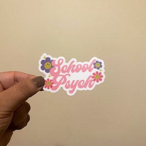 May include: A pink and white sticker with the text "School Psych" in a retro font. The text is surrounded by purple and pink flowers with yellow smiley faces.