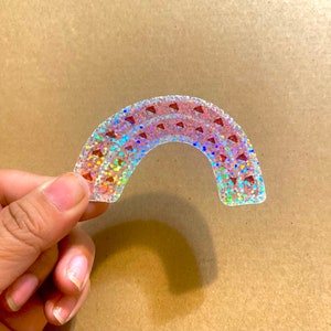 May include: A rainbow-colored sticker with a holographic finish and a pattern of red glitter hearts. The sticker is shaped like a rainbow arch.