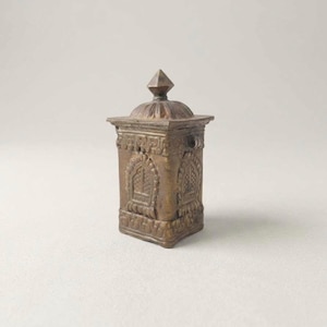 May include: A bronze-colored, ornate box with a pyramidal top. The box features detailed relief carvings of arched windows and decorative patterns. The box is rectangular with a square base and is likely a decorative item or container.