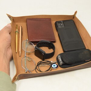 May include: A brown leather tray with a variety of items, including a phone, wallet, glasses, pens, a watch, and a necklace. The tray is made of high-quality leather and has a distressed look.