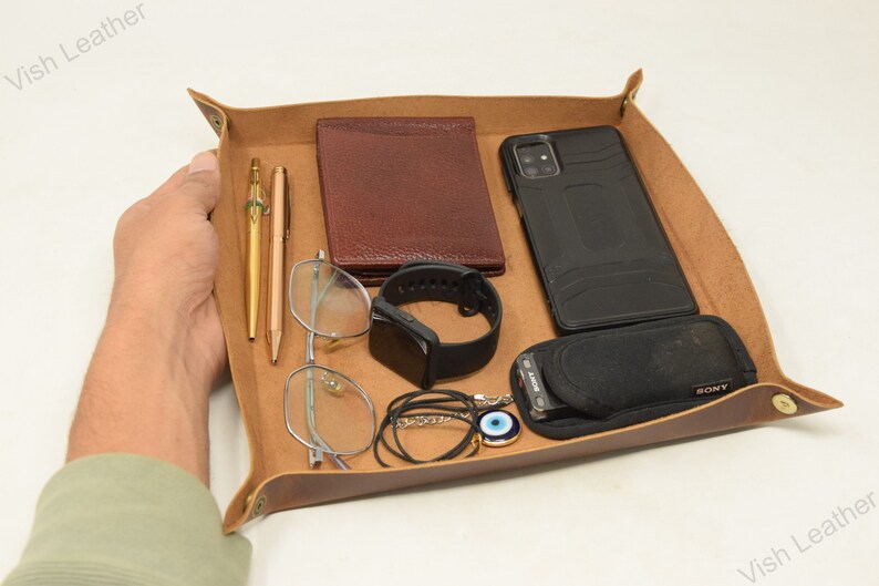 May include: A brown leather tray with a variety of items, including a phone, wallet, glasses, pens, a watch, and a necklace. The tray is made of high-quality leather and has a distressed look.