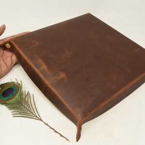 May include: A brown leather tray with a slightly distressed finish. The tray has a square shape with rounded corners and a raised edge. The tray is made of genuine leather and has a natural, rustic look.
