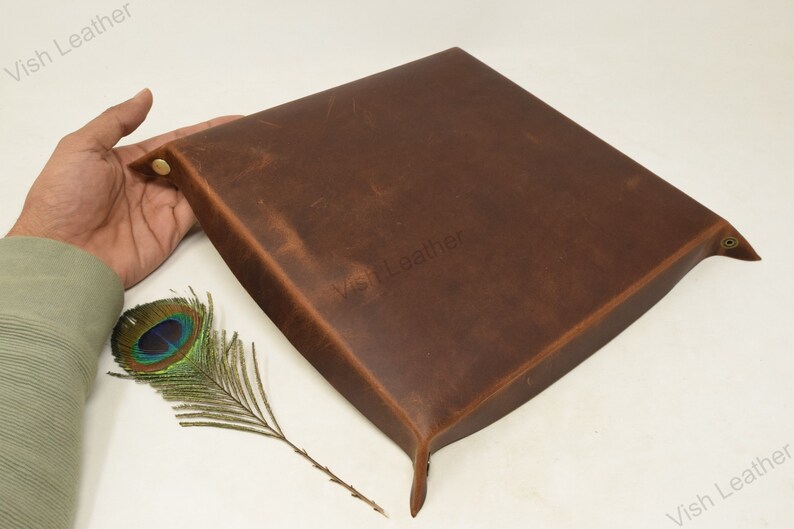 May include: A brown leather tray with a slightly distressed finish. The tray has a square shape with rounded corners and a raised edge. The tray is made of genuine leather and has a natural, rustic look.