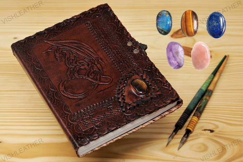 May include: A brown leather journal with a dragon design embossed on the cover. The journal has a tiger's eye stone inlay and is closed. Two calligraphy pens and five polished gemstones are on a wooden surface.