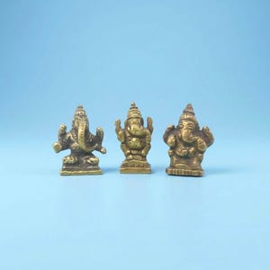 May include: Three small, detailed brass statues of the Hindu deity Ganesha are displayed against a light blue background. Each statue has a unique pose and intricate detailing, showcasing the elephant-headed god.