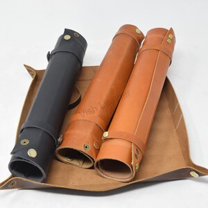 May include: Three leather rolls, one black and two brown, are displayed in a brown leather tray. The rolls have brass buckles and snaps.