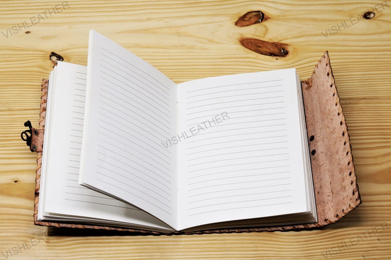 May include: A brown leather-bound journal with lined pages is open on a wooden surface. The journal has a decorative stitching detail on the cover.
