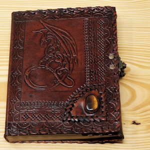 May include: A brown leather journal with a dragon design embossed on the front cover. The journal has a clasp closure and a tiger's eye stone inlay.