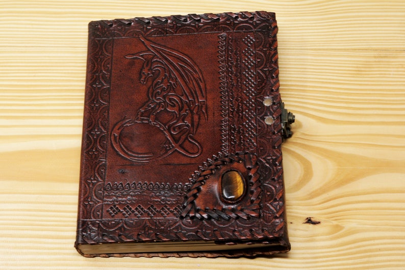 May include: A brown leather journal with a dragon design embossed on the front cover. The journal has a clasp closure and a tiger's eye stone inlay.