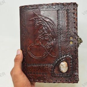 May include: A brown leather journal with an embossed dragon design and a decorative stone inlay. The journal has a metal clasp and is stitched with brown thread.