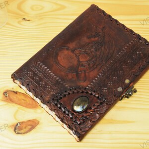 May include: A brown leather journal with a detailed embossed design of a dragon and Celtic knotwork. The journal has a decorative metal clasp and a gemstone inlay.