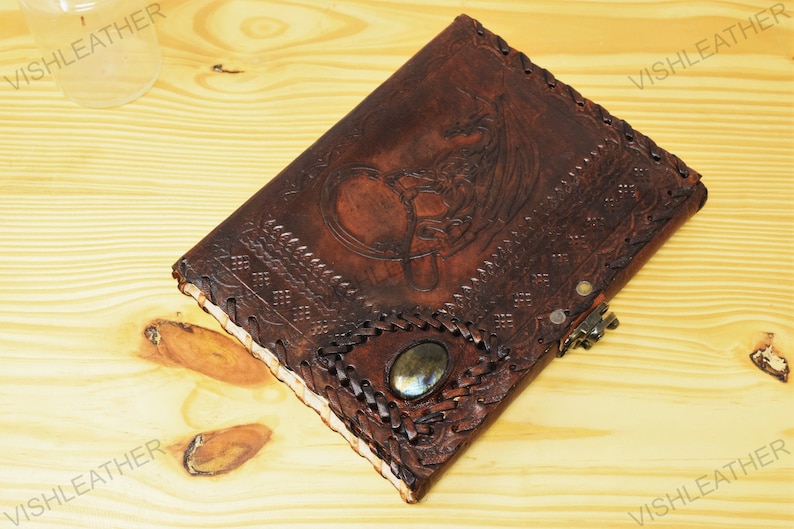 May include: A brown leather journal with a detailed embossed design of a dragon and Celtic knotwork. The journal has a decorative metal clasp and a gemstone inlay.