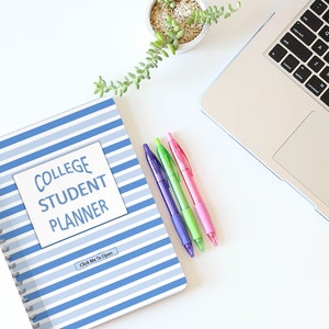 May include: A blue and white striped college student planner with the words "College Student Planner" on the front. The planner is on a white desk with a laptop, a potted plant, and three pens.