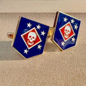 Op de afbeelding: Een paar vijfhoekige manchetknopen in goudkleur met een donkerblauwe emaille achtergrond. Elke manchetknop toont een centrale rode ruit met een witte schedel, omringd door vijf kleine witte sterren in een cirkelvormig patroon op het blauwe veld.