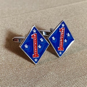 May include: A pair of silver cufflinks with a blue and red enamel design. The design features a diamond shape with a white number one and the words "Guadalcanal" in red.