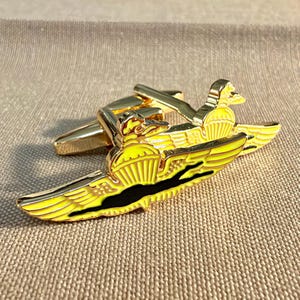 May include: A gold-toned metal cufflink featuring a winged parachute emblem with bright yellow enamel fill and a black horizontal stripe across the center. The design includes a stylized bird head at the top, with a hinged toggle backing.